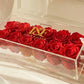 Red roses in a clear box on a marble surface with white flowers and a mirror in the background.