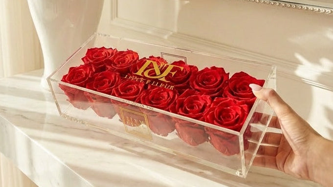 Red roses in a clear box on a marble surface with white flowers and a mirror in the background.